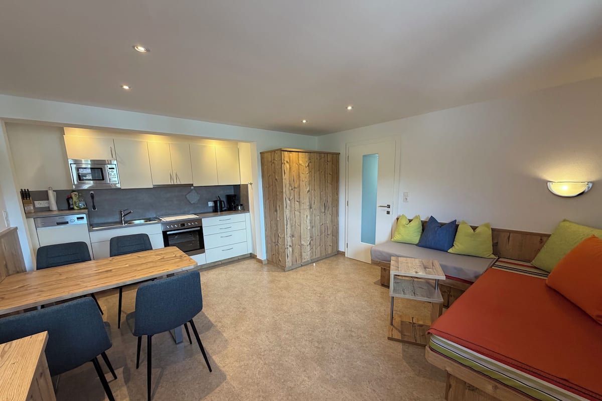 A modern kitchen with built-in appliances, a dining area with a wooden table and chairs, and a cozy seating area with a sofa and cushions.