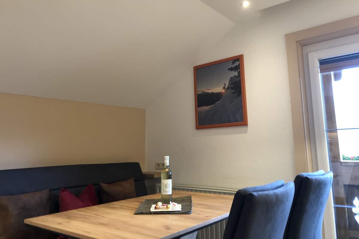 A dining area with a wooden table, a bottle of wine, and a plate of food. There is a painting on the wall and a window with curtains.