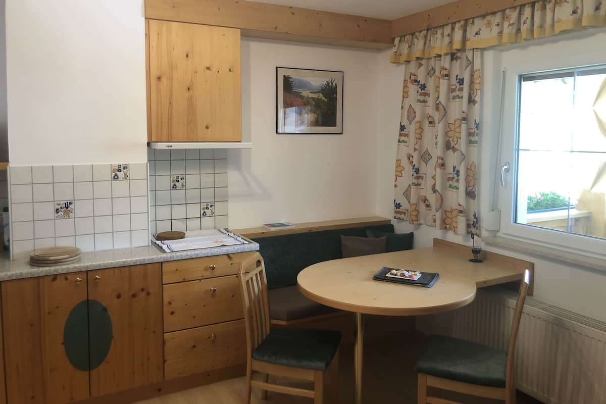 A small kitchen with wooden cabinets, a tiled backsplash, a round table with chairs, and a window with curtains.