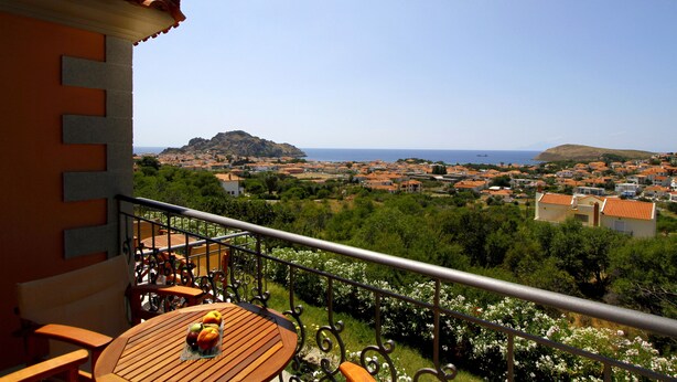 A balcony with a wooden table and chairs, overlooking a coastal town with houses and greenery. - Amygdalies Apartments (Lemnos)
