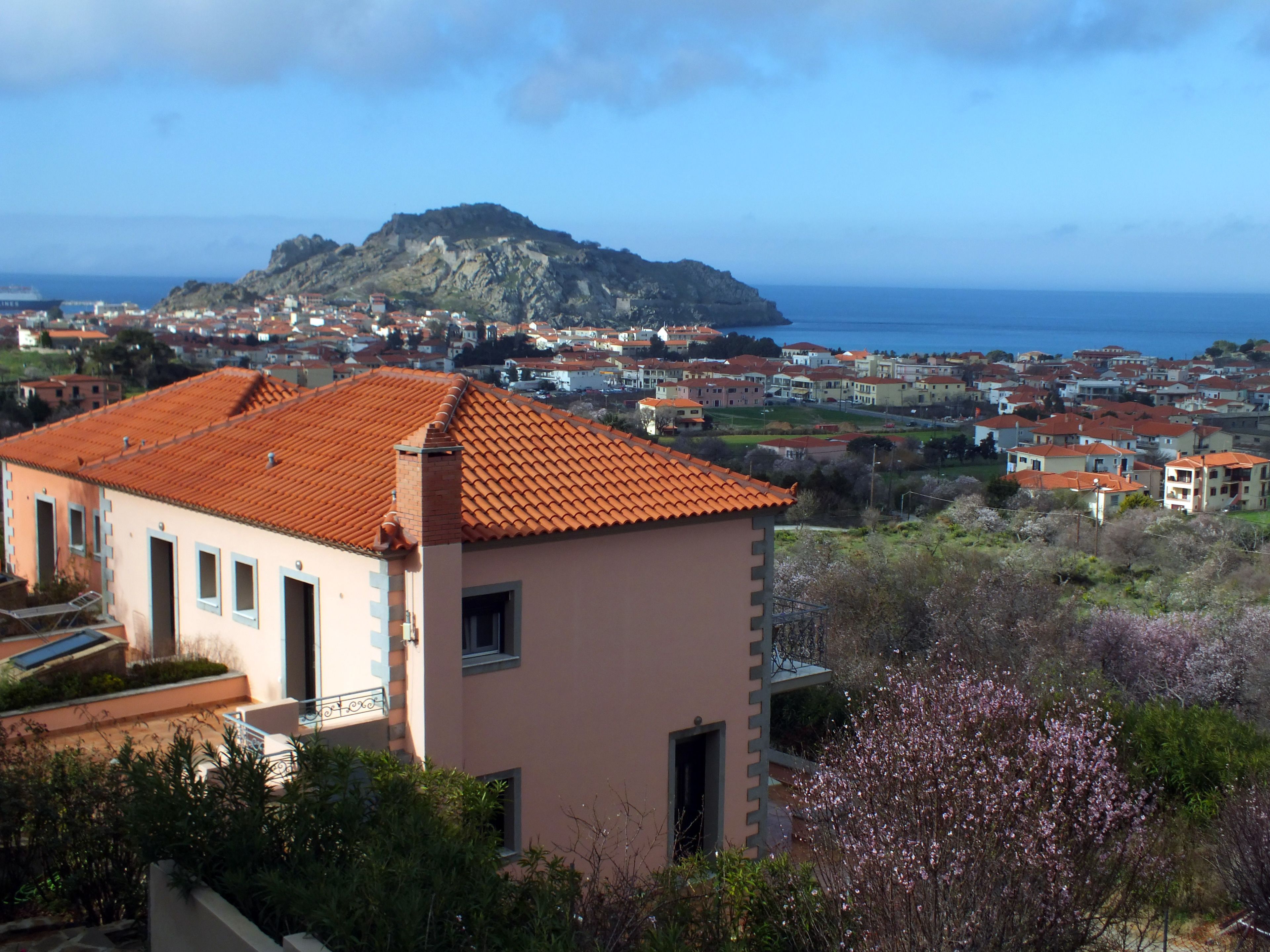 A coastal town with a prominent hill in the background, a pink building with a red-tiled roof, and a view of the sea.