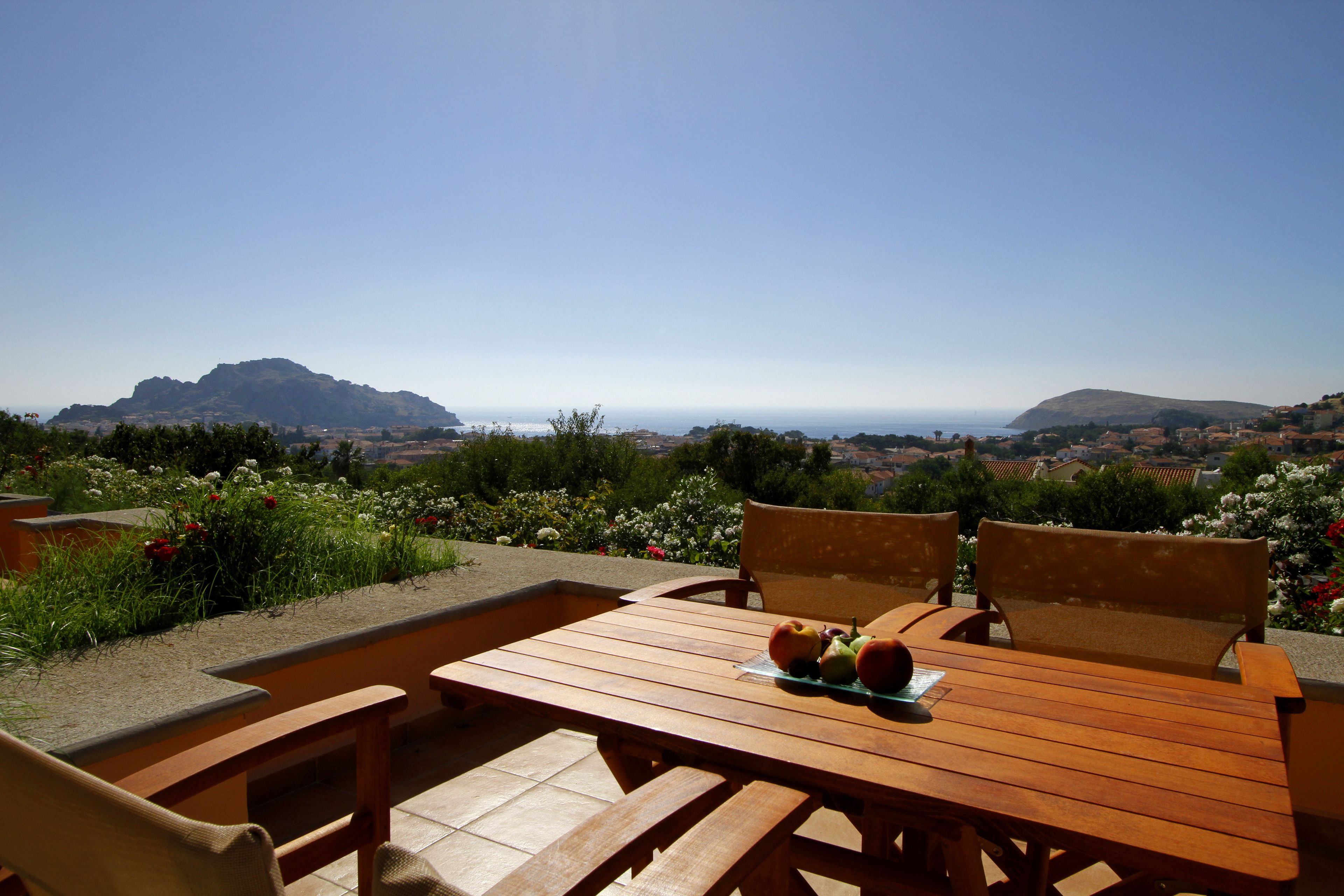 Una mesa de madera con dos sillas y un plato de fruta, con vistas a un paisaje pintoresco con montañas y el mar.