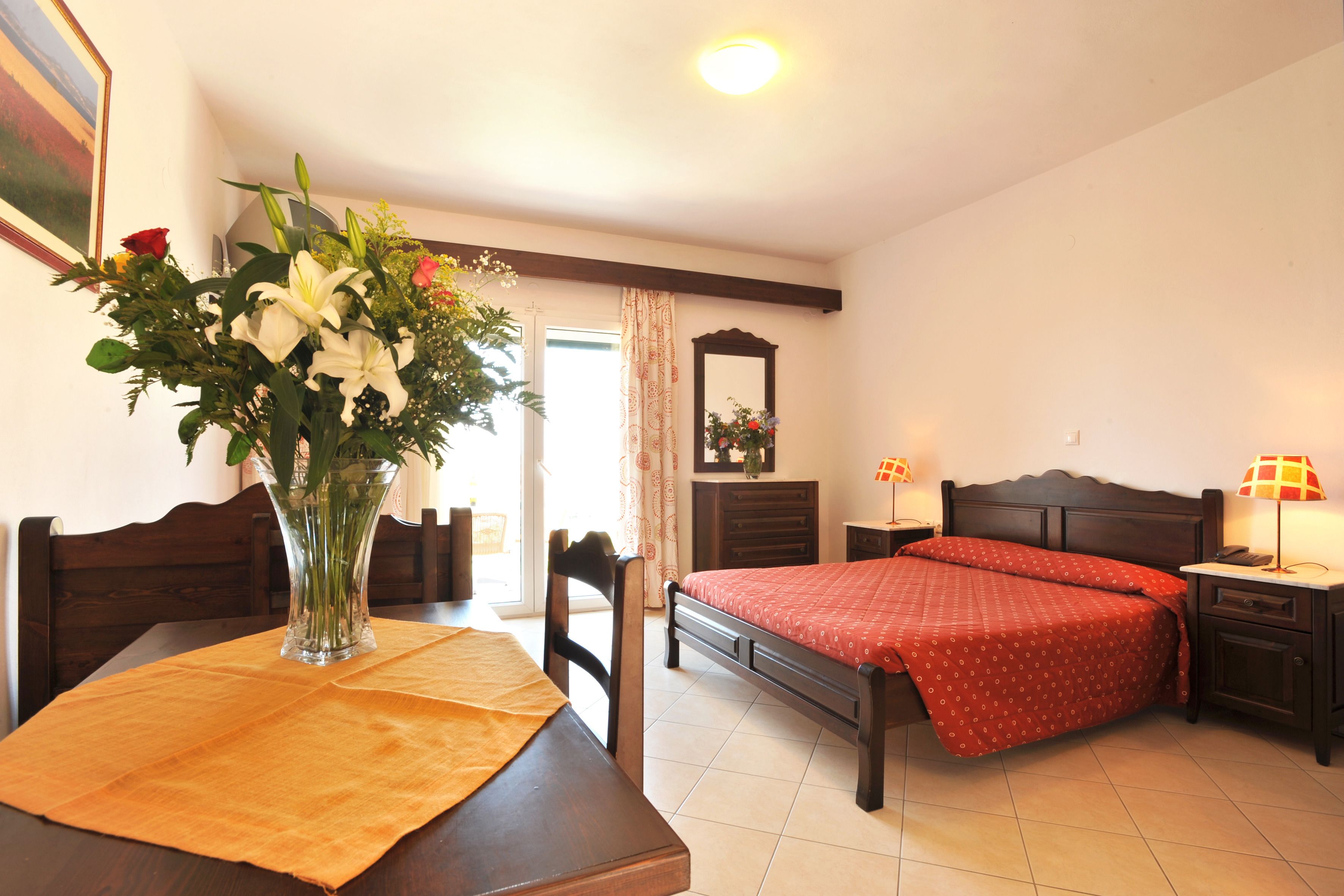 A bedroom with a bed, bedside table, nightstand, and a vase of flowers on a table.