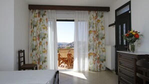 A bedroom with a bed, a wooden chair, a dresser, and a view of a balcony with a table and chairs. - Amygdalies Apartments (Lemnos)