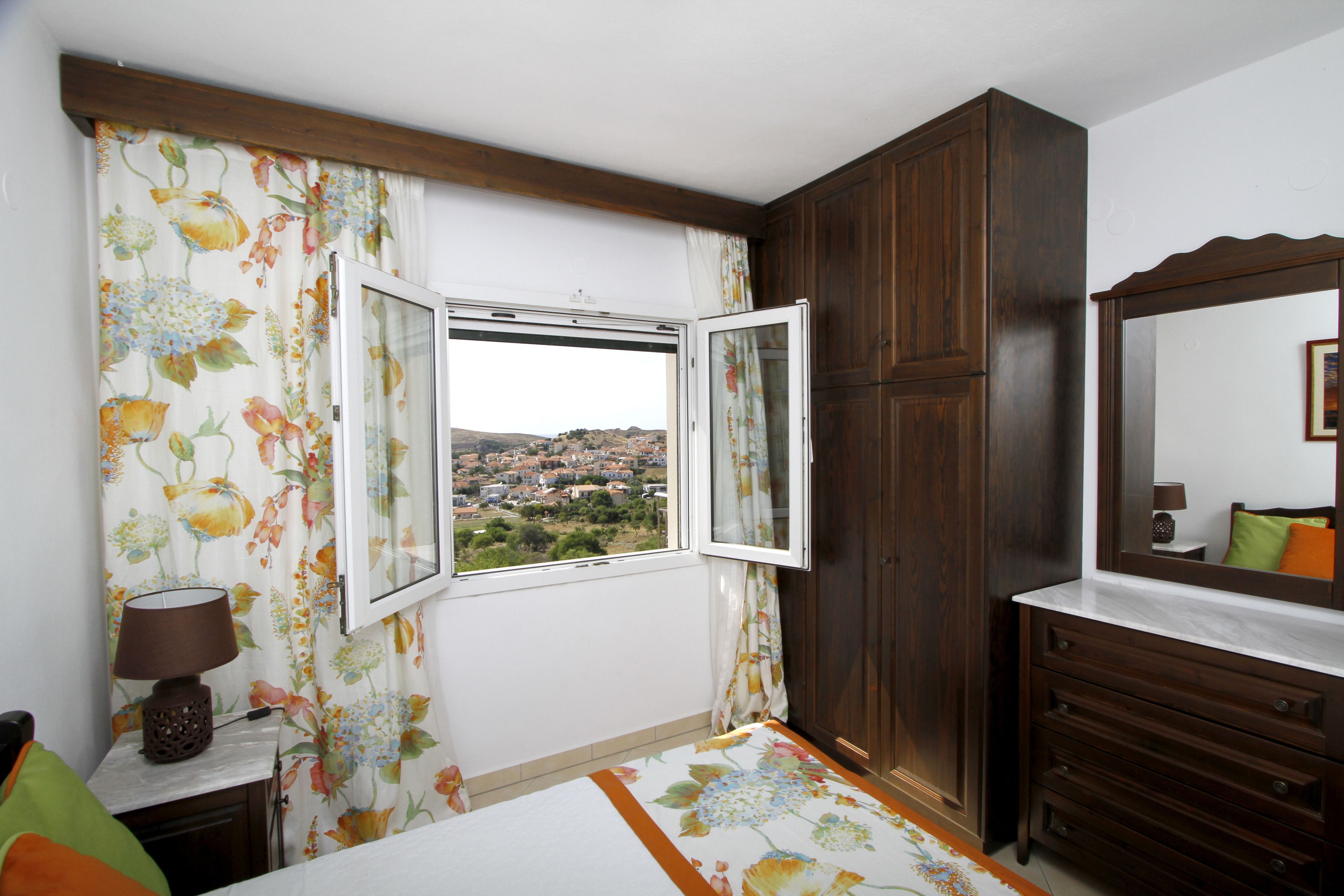 A bedroom with a large window, wooden wardrobe, and a bedside table with a lamp.