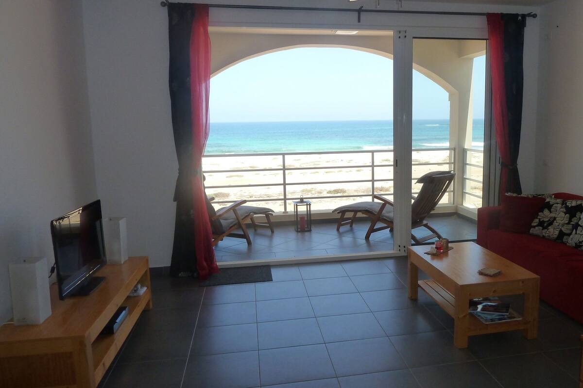 Ein Wohnzimmer mit Fernseher, einem roten Sofa, einem Couchtisch aus Holz und Blick auf Strand und Meer durch eine offene Tür.