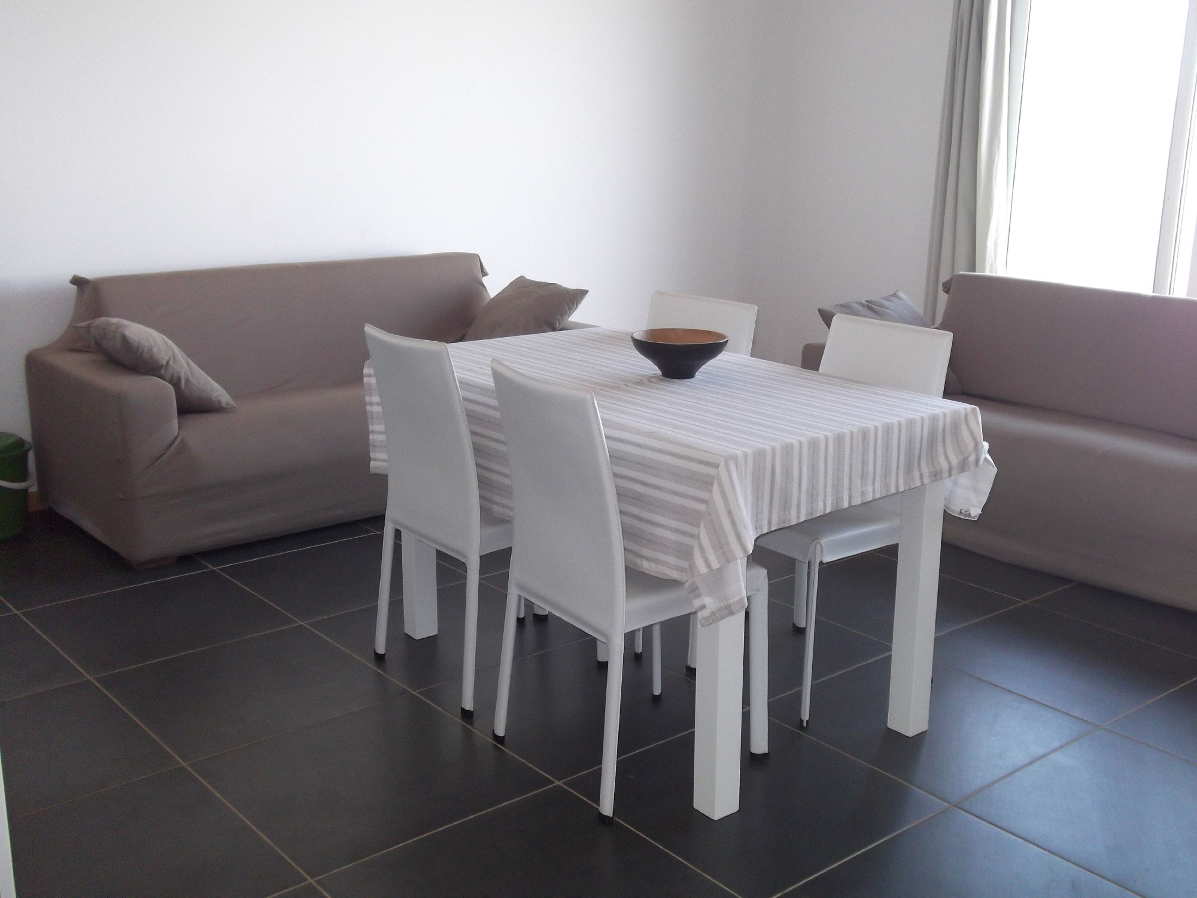 A modern living room with a dining table, white chairs, a sofa, and a large window.