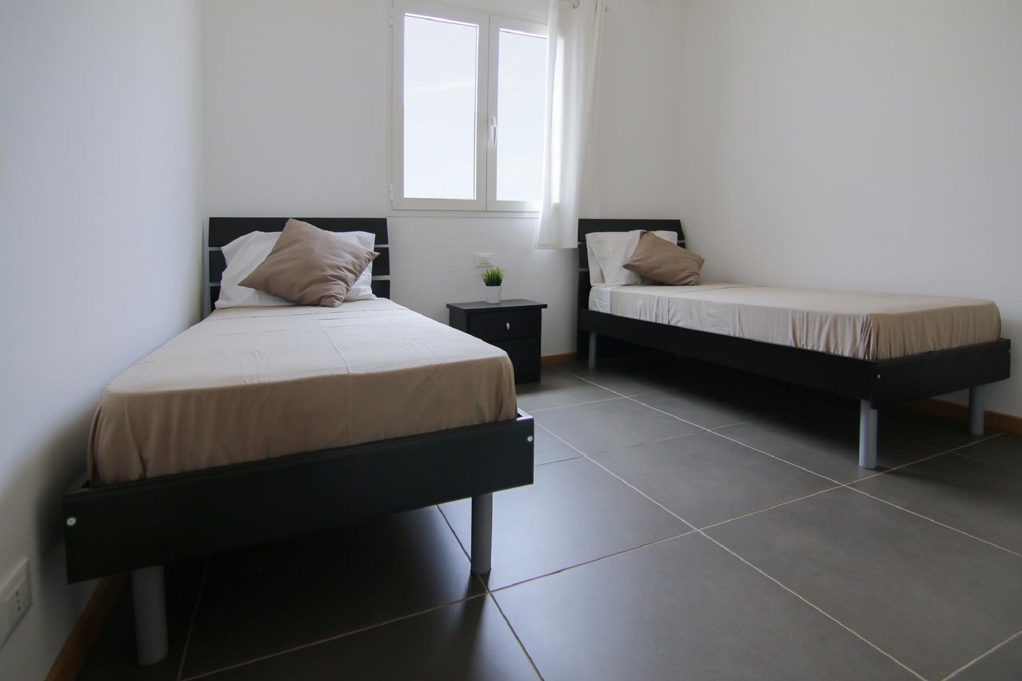 A room with two single beds, a small bedside table with a lamp, and a window with curtains.