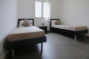 A room with two single beds, a small bedside table with a lamp, and a window with curtains. - Ca Jasmine (Boa Vista)