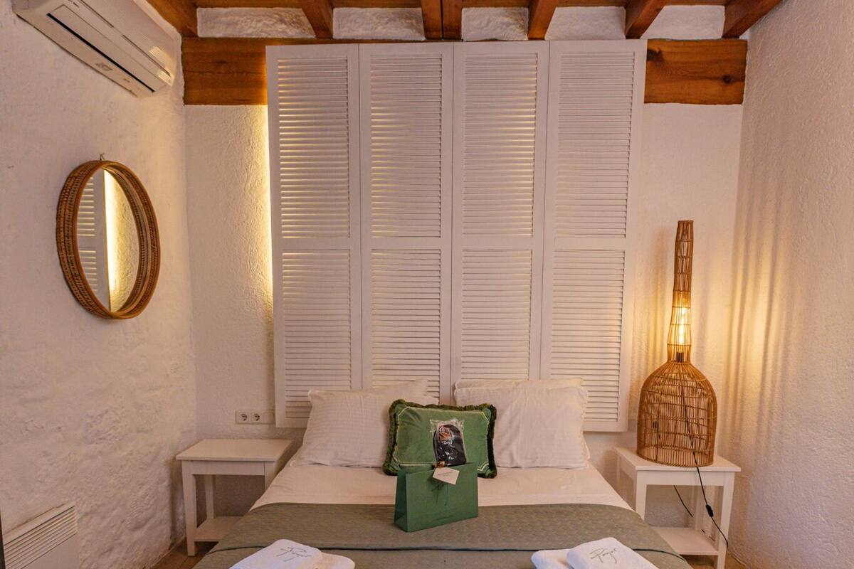 Ein Schlafzimmer mit einem großen Bett, weißen Wänden, Holzbalken an der Decke, einem Nachttisch, einem Spiegel und einer Lampe.
