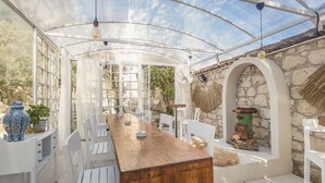 Un espace de restauration extérieur abrité, doté d’une longue table en bois et de chaises blanches, entouré de murs en pierre et surmonté d’un plafond en verre.