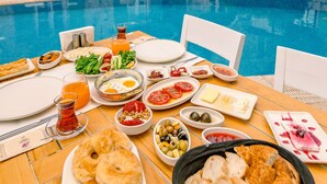 Une table à manger dressée avec une variété de plats et de boissons, placée devant une piscine et offrant une vue sur un bâtiment en pierre aux volets verts.