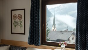 Een kamer met een bed, een raam met gordijnen, een ingelijste botanische print en een vaas met bloemen.