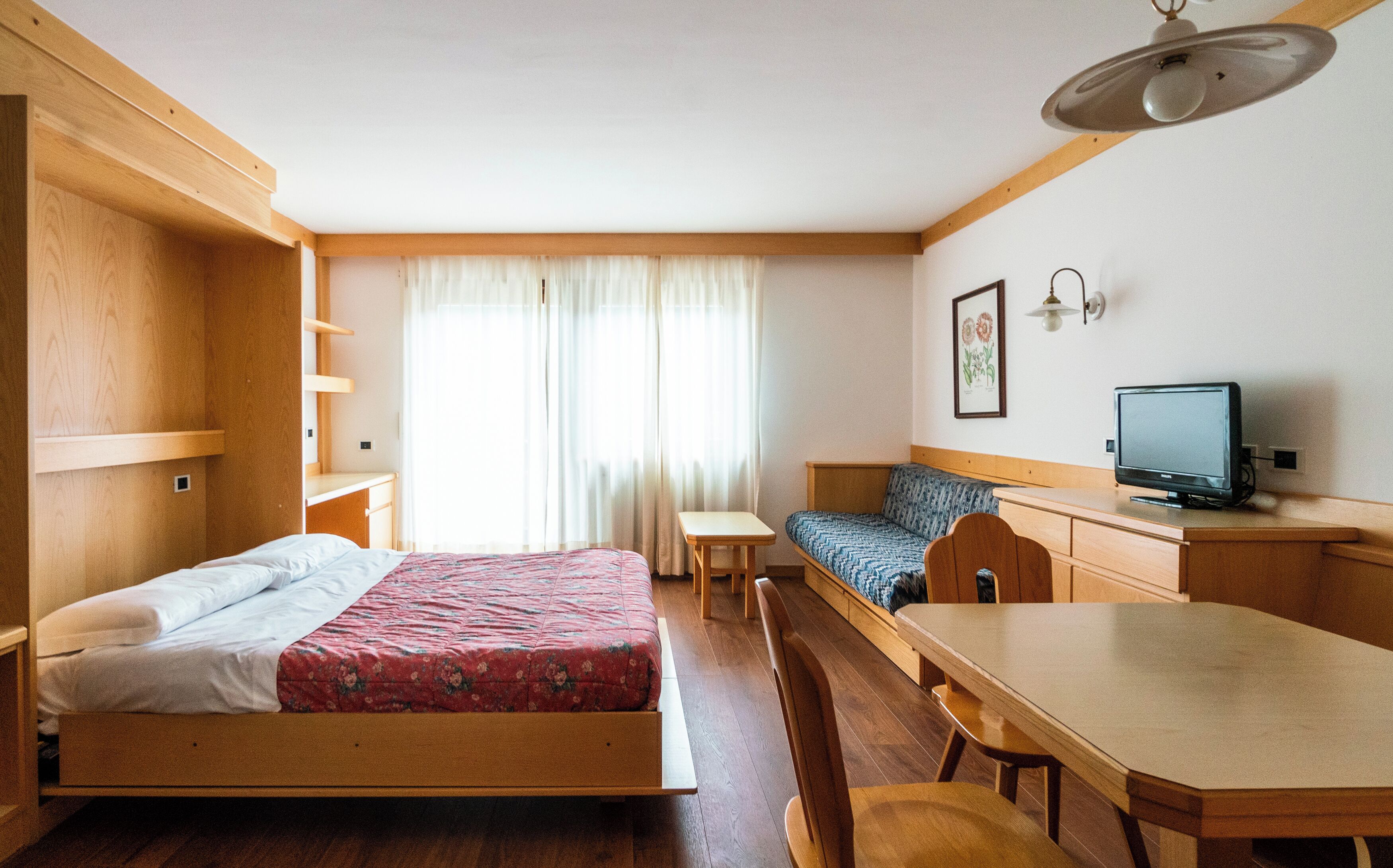 A compact room with a bed, a desk, a chair, a small table, a sofa, and a television.