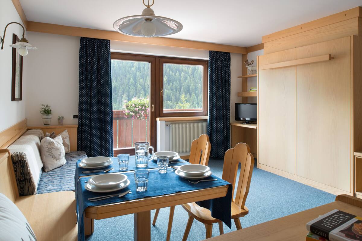 Een eetruimte met een tafel gedekt voor vier personen, houten stoelen, een blauwe tafelkleed en uitzicht op een bos door een raam.
