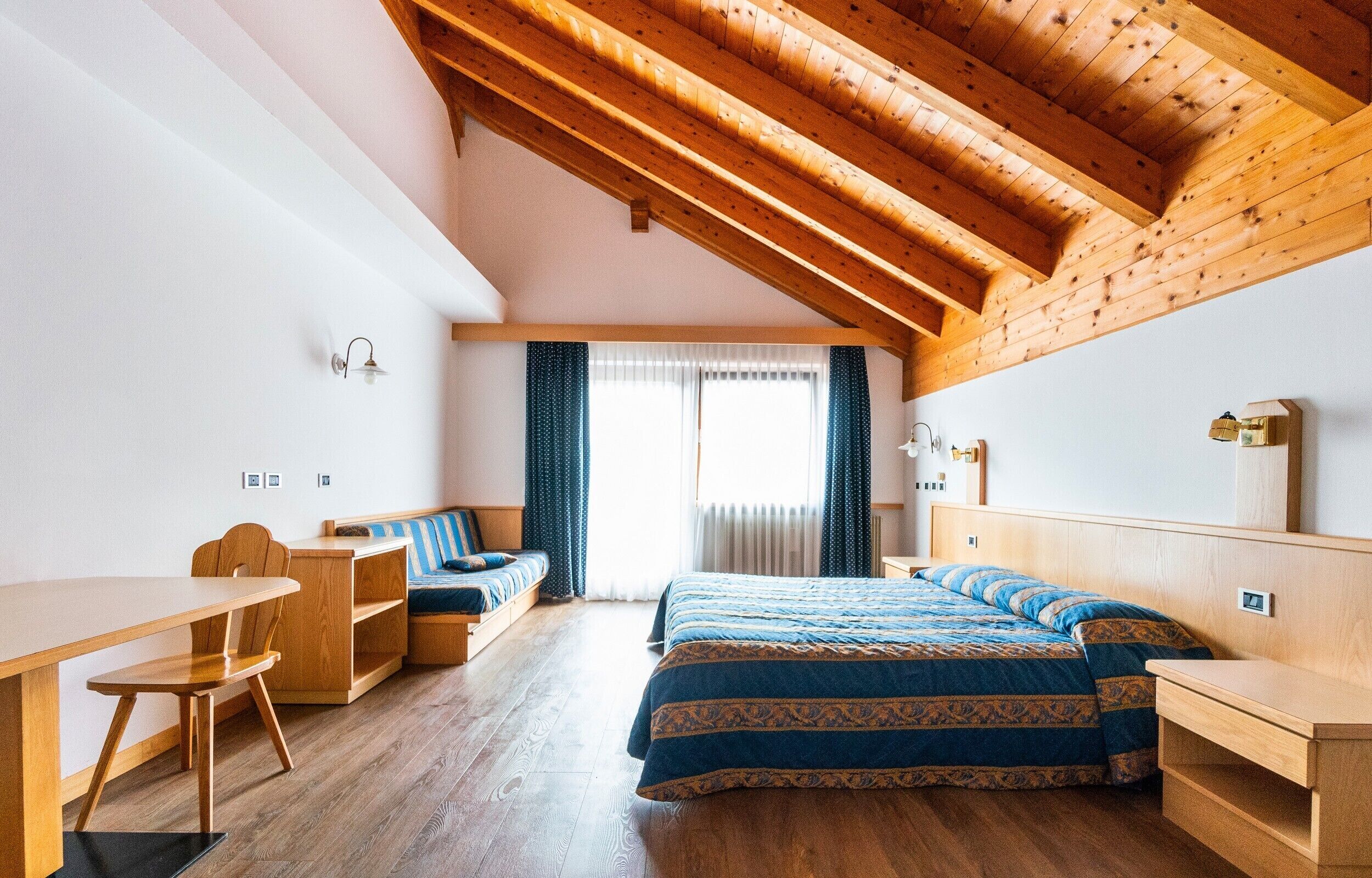 A room with a sloped ceiling, wooden beams, a bed with patterned bedding, a desk, a chair, and a window with curtains.