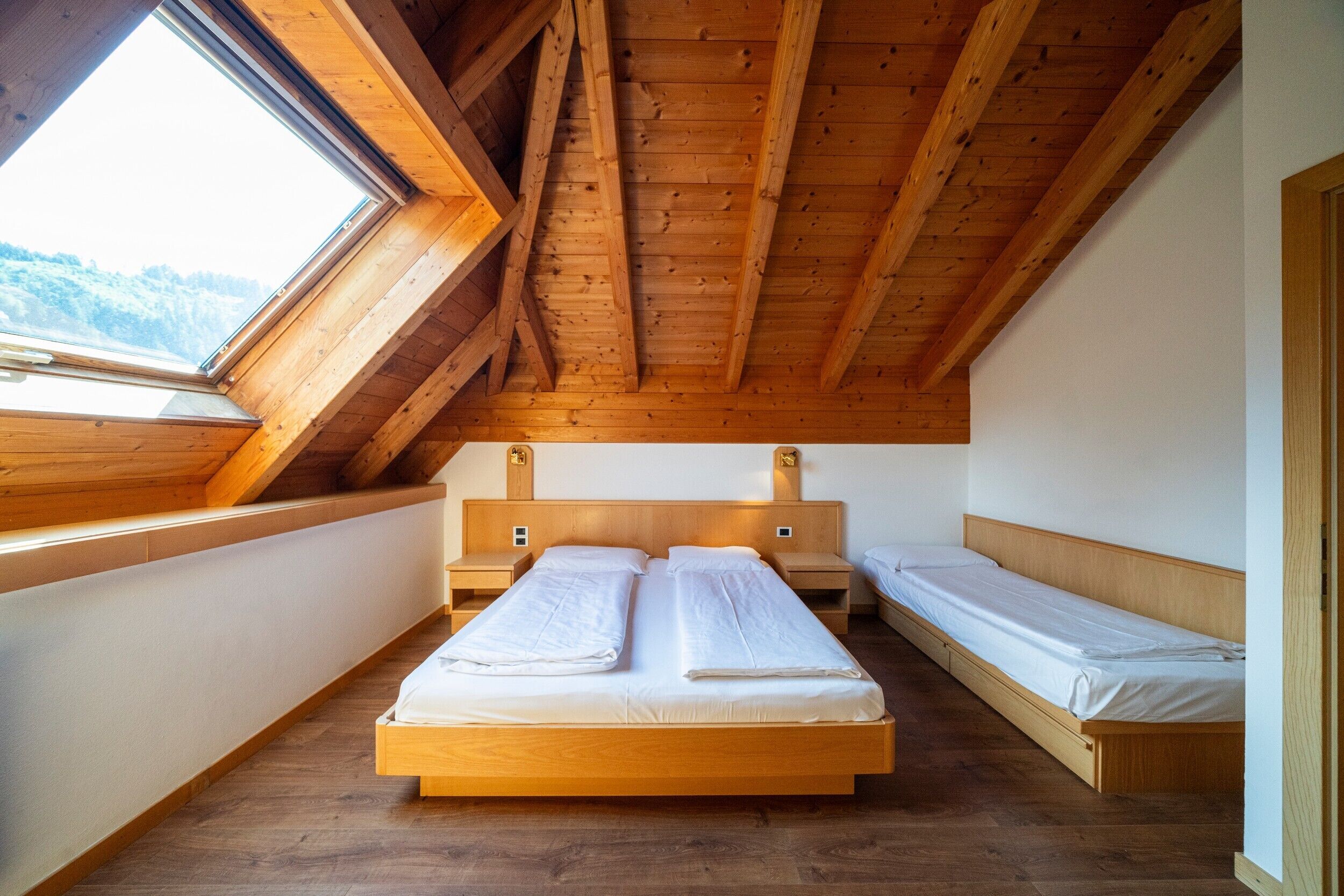 A room with a sloped ceiling, wooden beams, a large skylight, and two single beds with wooden headboards.