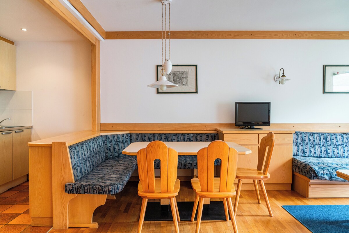 Une cuisine moderne avec des meubles en bois, un coin repas avec un banc et des chaises, et un téléviseur posé sur une table en bois.