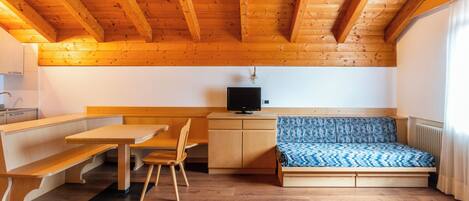 A compact room with a wooden ceiling, a bench, a table, a chair, a small desk with a computer, and a bed with patterned bedding.