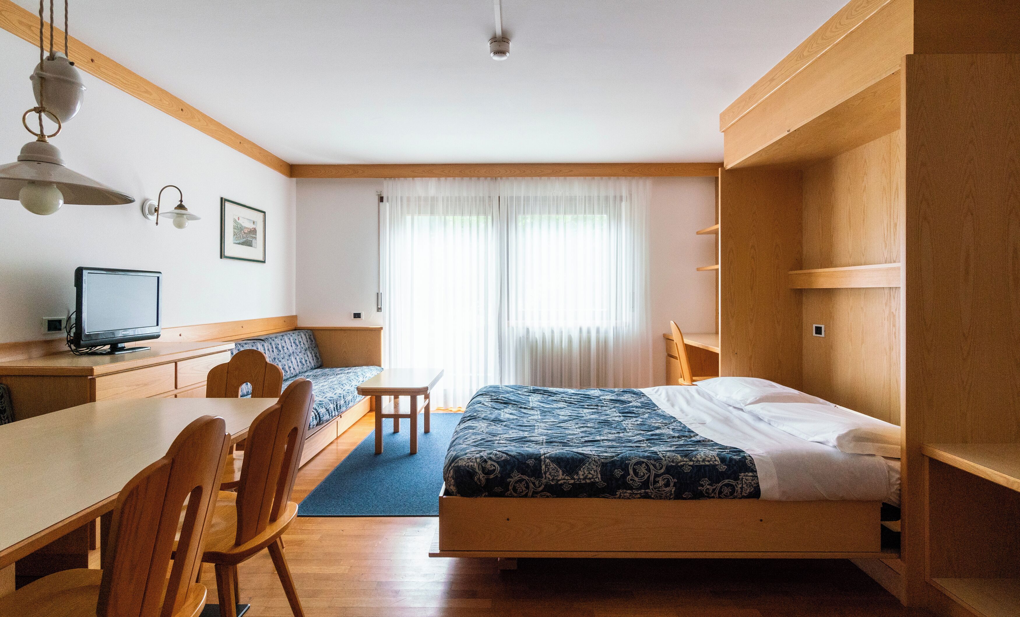A hotel room with a bed, a desk with a computer, a chair, and a window with curtains.
