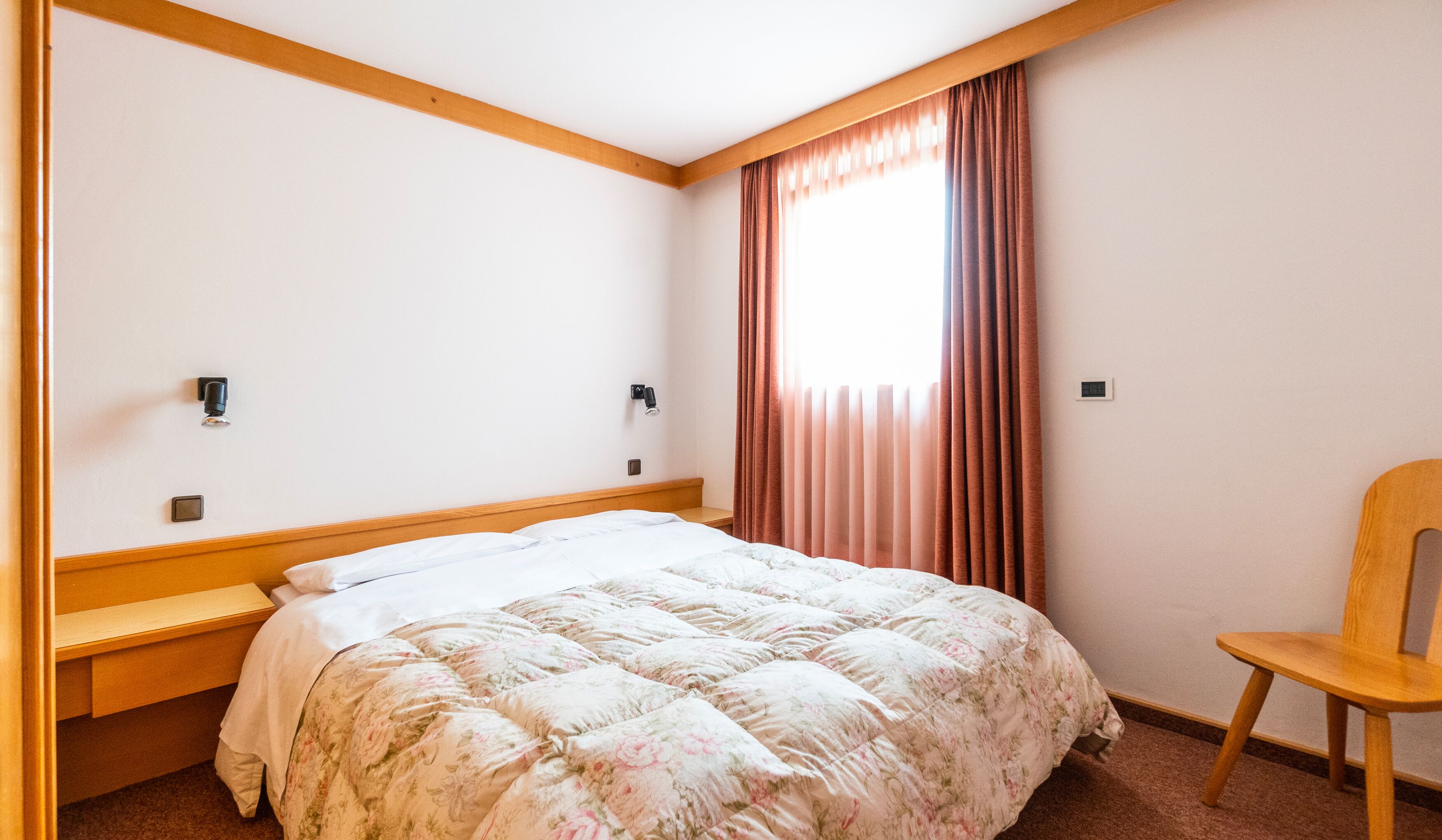 A bedroom with a bed, a wooden chair, a window with curtains, and a wall-mounted light fixture.