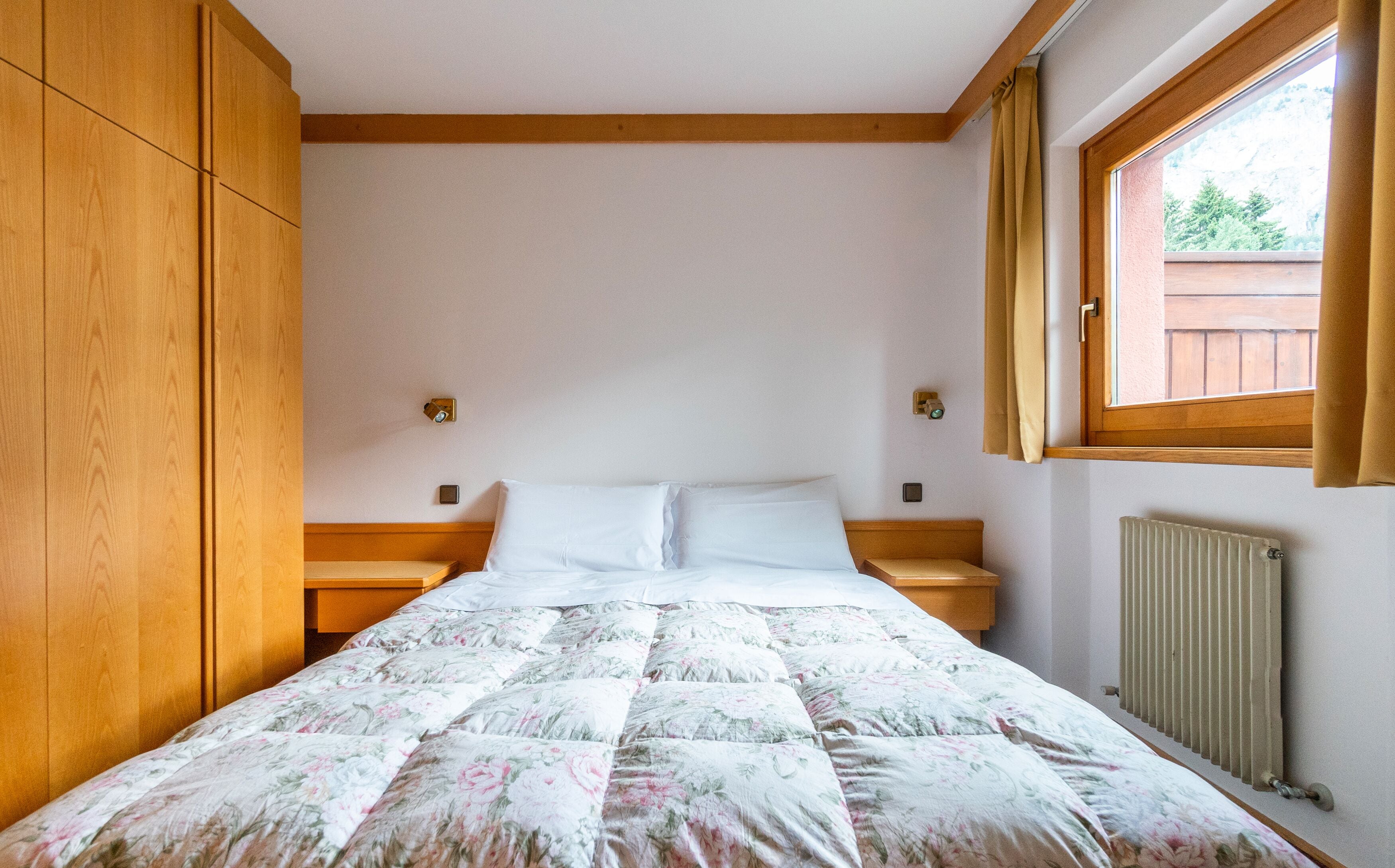 Un dormitorio con un armario de madera, una cama con ropa de cama floral, dos mesitas de noche y una ventana con cortinas.
