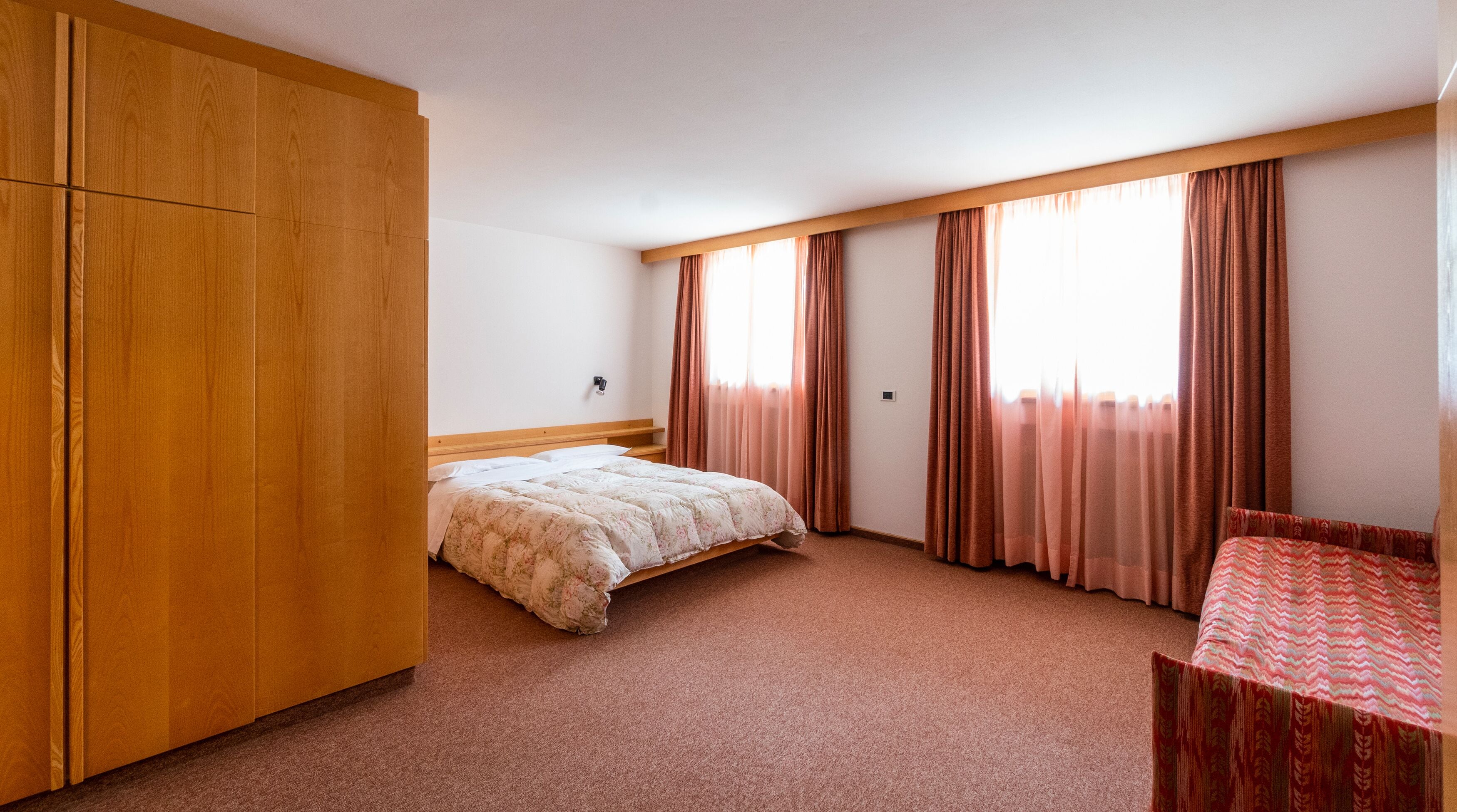 A bedroom with a bed, a wooden wardrobe, a desk, and large windows with curtains.