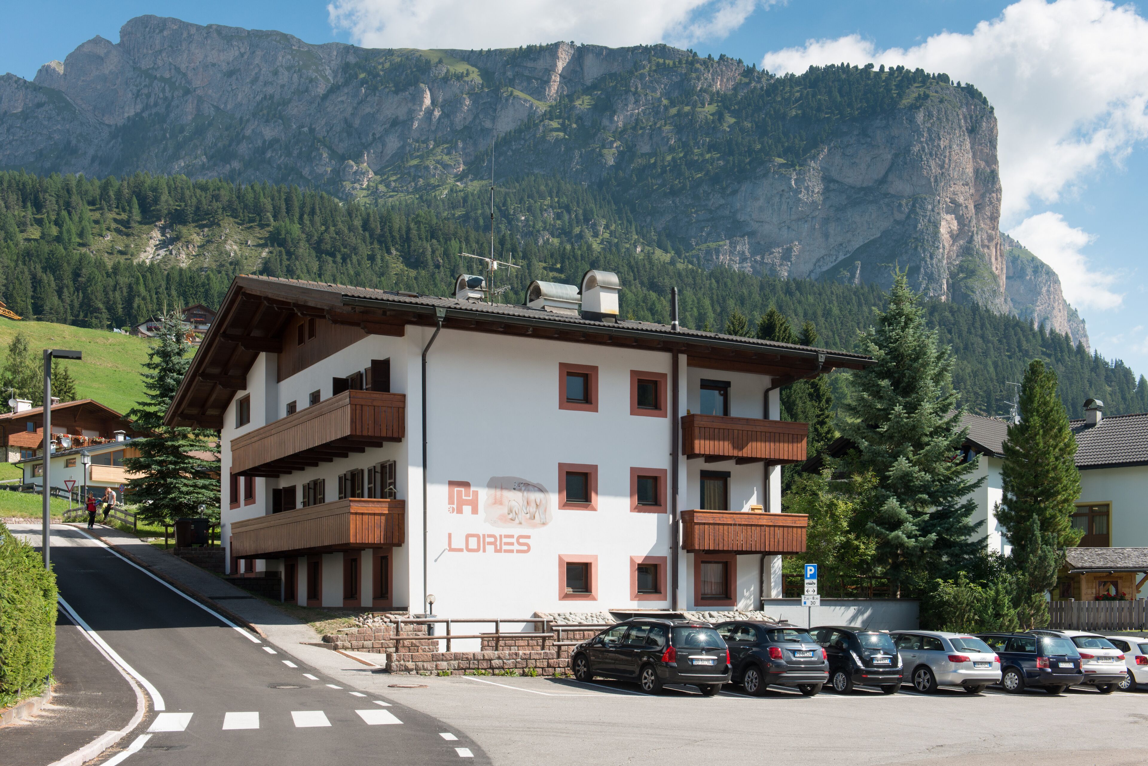 A multi-story hotel with balconies, a parking lot, and a mountainous backdrop.