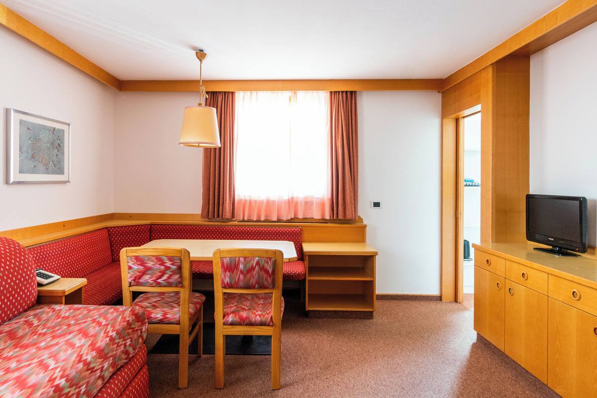 A cozy room with a red sofa, wooden chairs, a small table, a television, and a window with curtains.