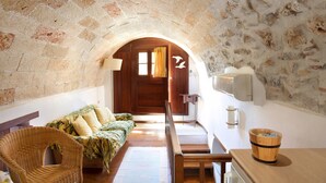 Ein gemütlicher Raum mit einem Steinbogen, einer hölzernen Bank, einem Korbstuhl, einer Couch mit gemusterten Kissen und einem Holztisch mit einem Eimer.