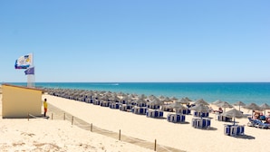A beach with rows of blue beach chairs and umbrellas, a flag, and a small building. - Dunas Douradas Beach Club (Loulé)