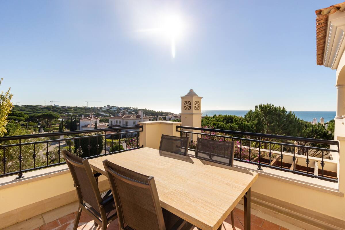 A balcony with a table and chairs, overlooking a coastal view with buildings and trees.