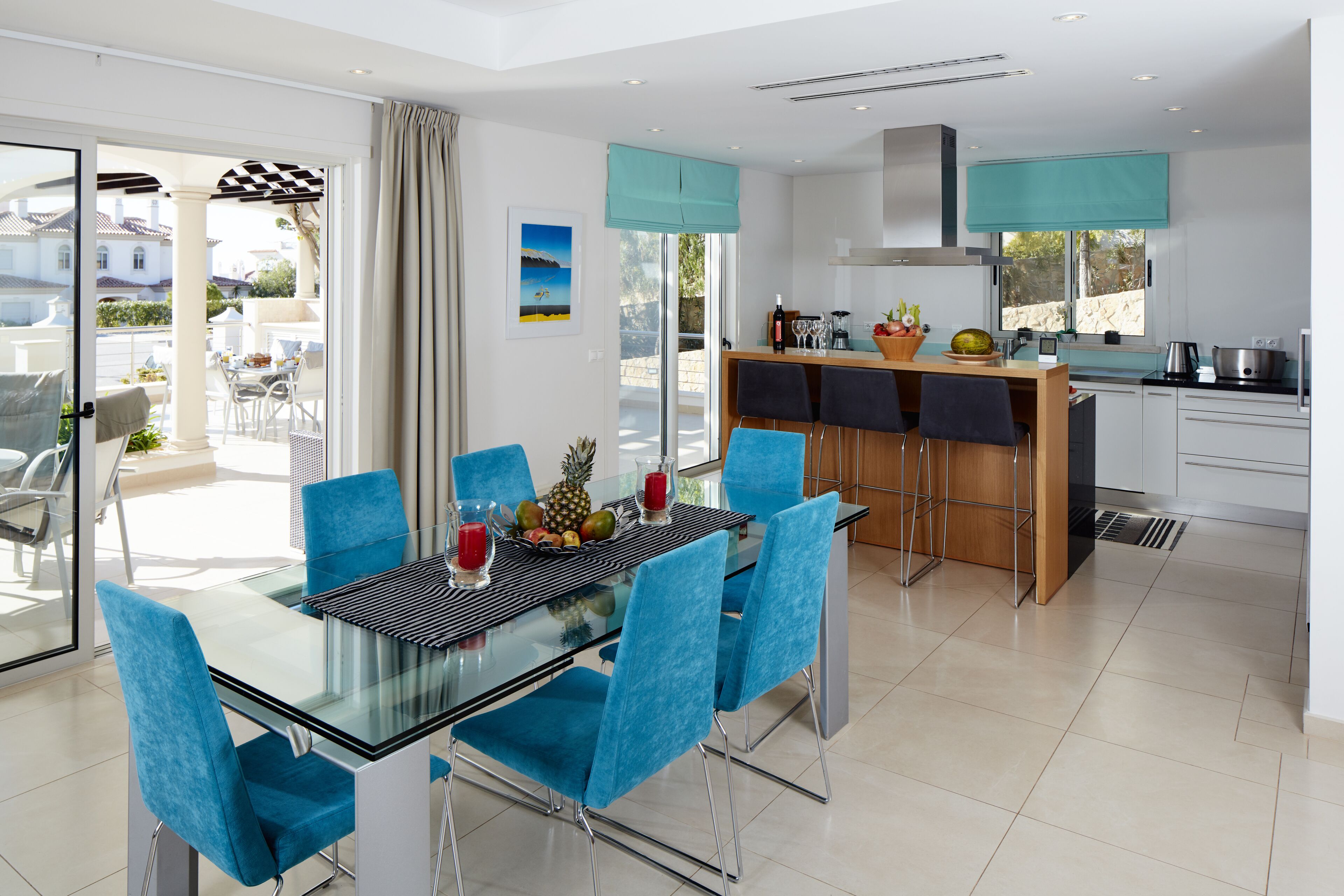 Cocina moderna con comedor, que incluye una mesa de vidrio y sillas azules, una isla de cocina y vistas al exterior.