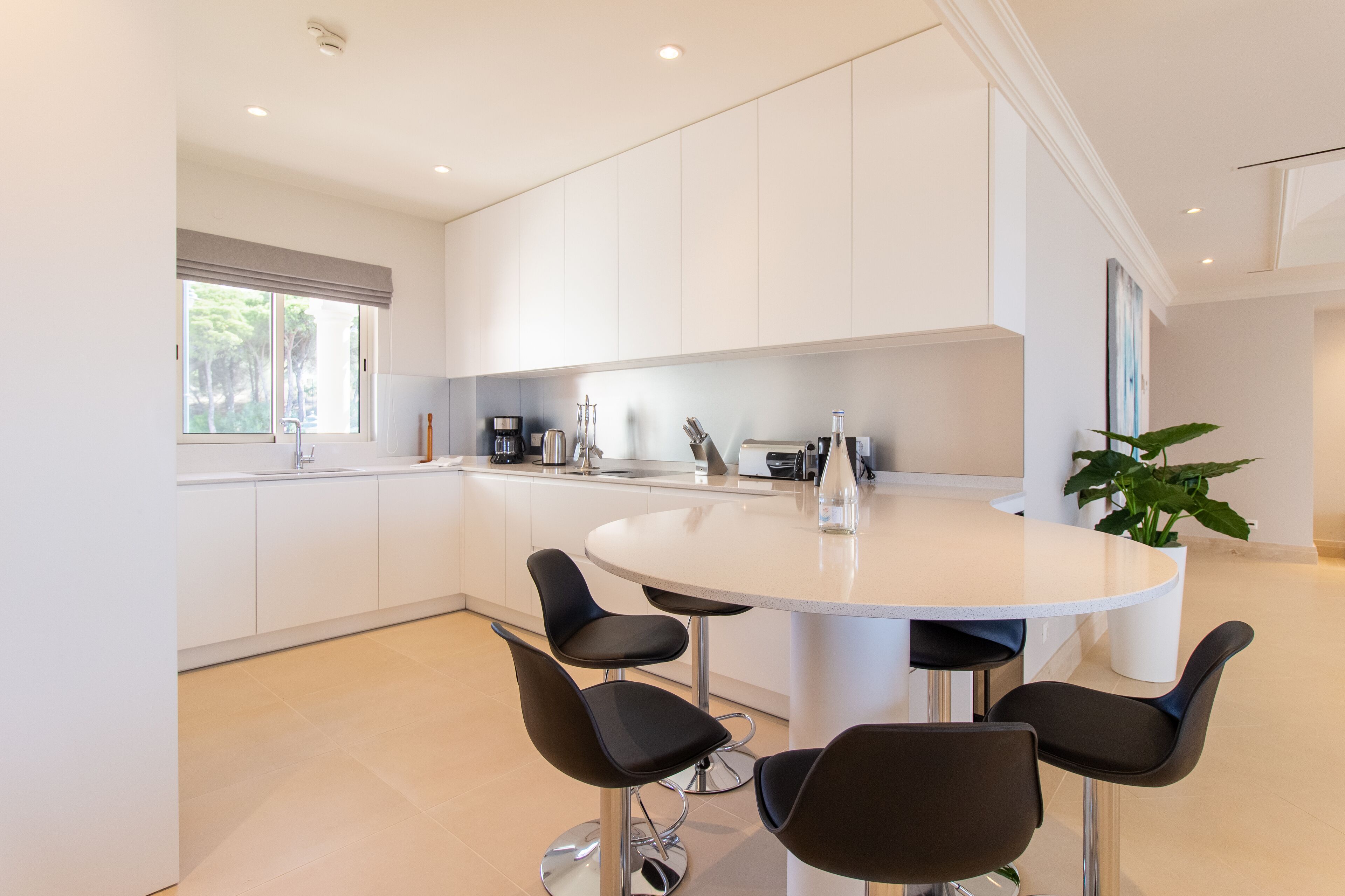 A modern kitchen with white cabinets, a round island with bar stools, and a large window.