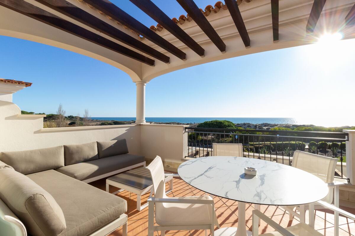 A terrace with a white sofa, a round table, and chairs, overlooking a scenic view of the sea.