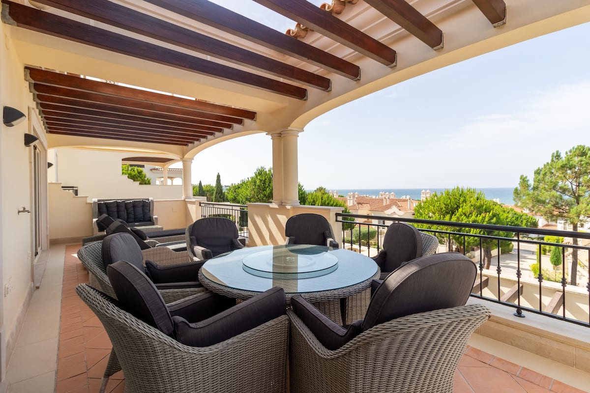 A covered outdoor seating area with wicker furniture and a glass table, overlooking a scenic view.