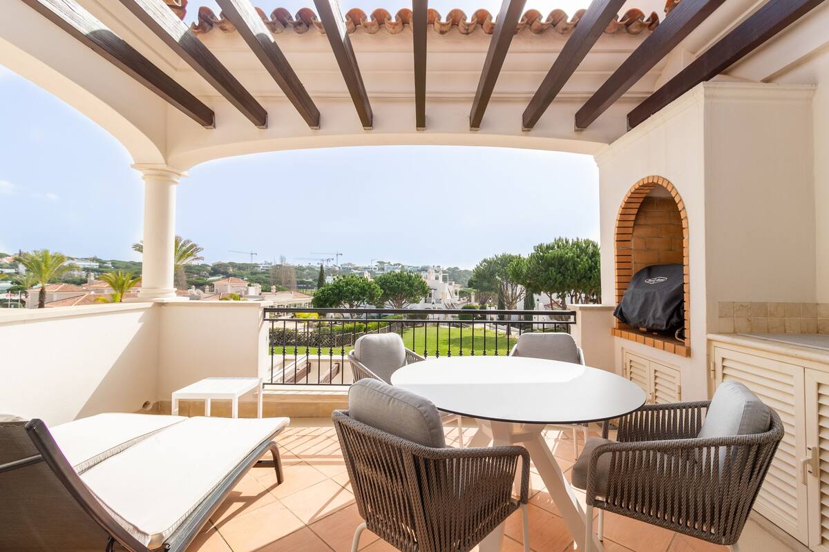 A terrace with a white table and wicker chairs, a brick oven, and a view of the cityscape.