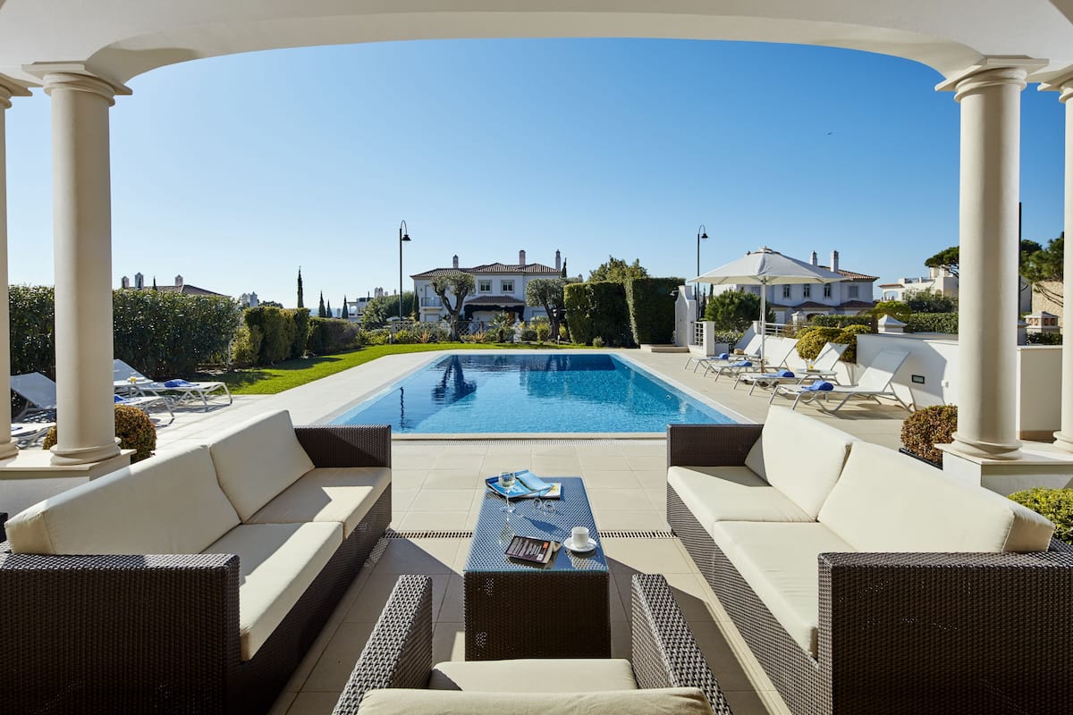 An outdoor pool area with lounge chairs and a sofa, surrounded by white columns and a clear blue sky.