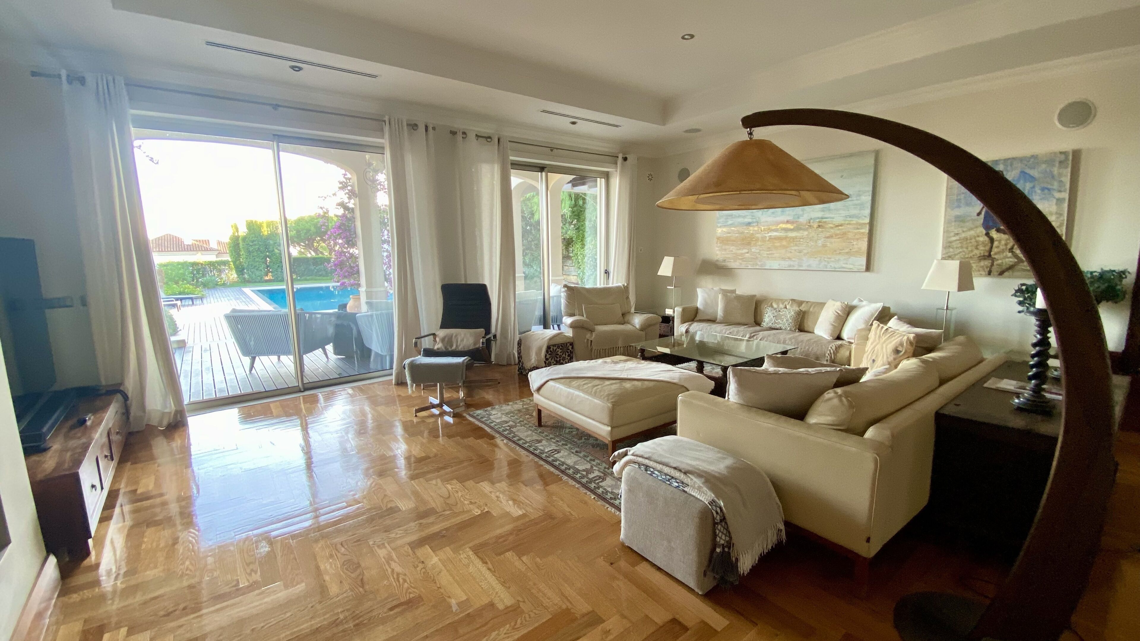 A spacious living room with a large sofa set, a coffee table, and a dining table. There is a sliding glass door leading to an outdoor area with a pool.