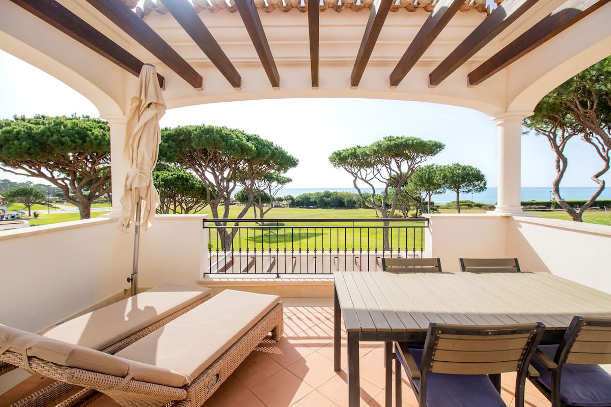 A covered outdoor seating area with a table and chairs, overlooking a green landscape and the sea.
