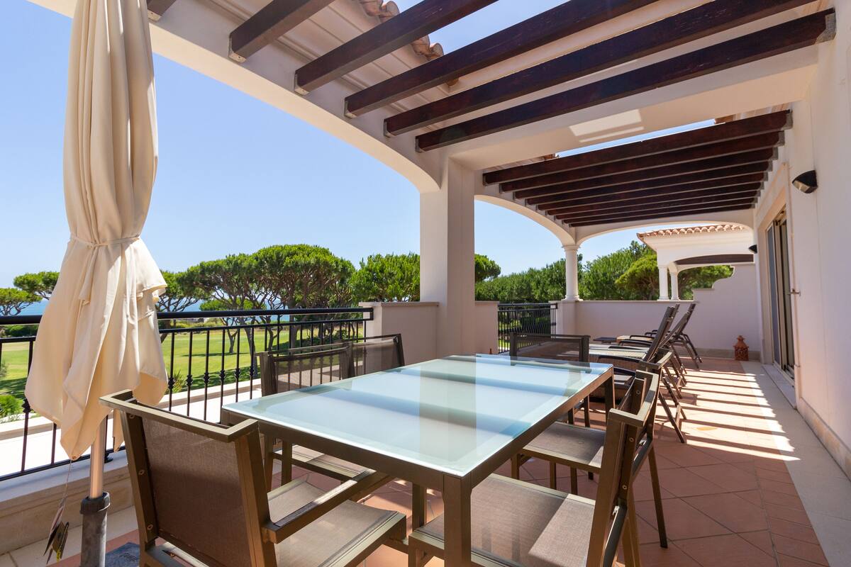An outdoor patio with a glass-top table and chairs, surrounded by a white wall and a pergola.