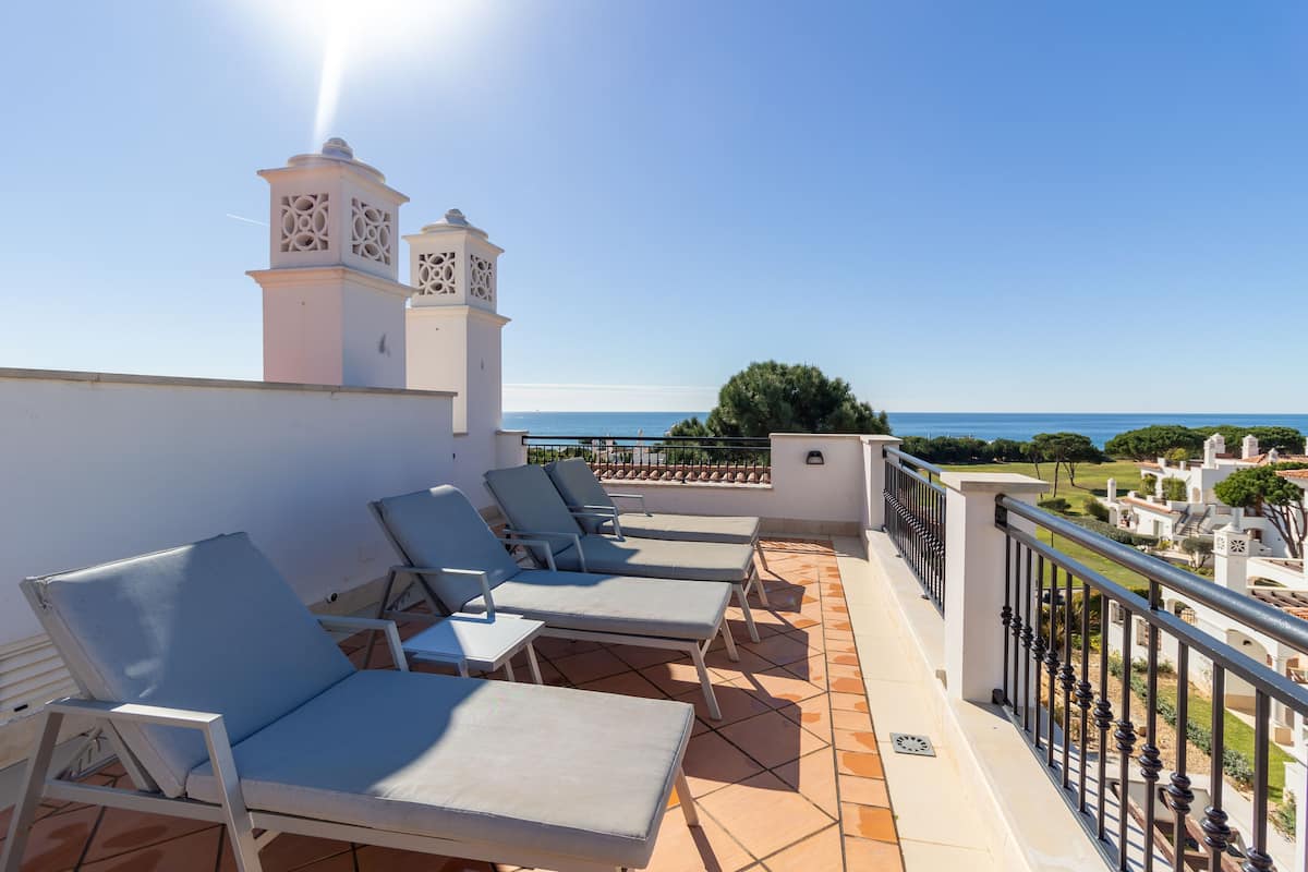 A balcony with four lounge chairs, a white wall, and a view of the sea.