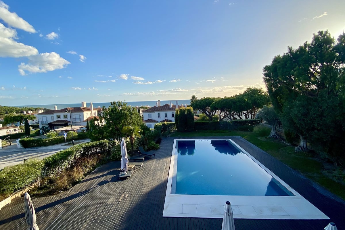 A swimming pool with a wooden deck, surrounded by greenery and a view of the sea and buildings.