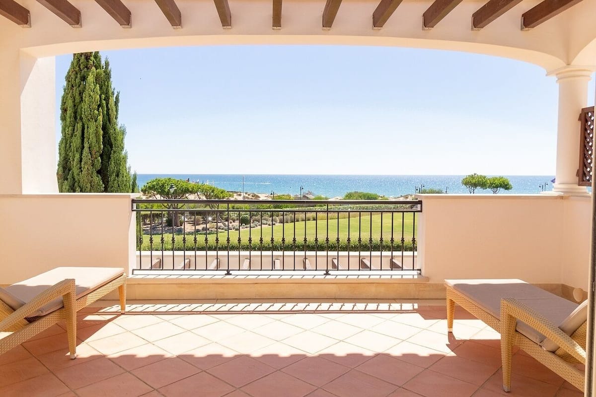 A balcony with two wicker lounge chairs, a tiled floor, and a view of the sea and greenery.
