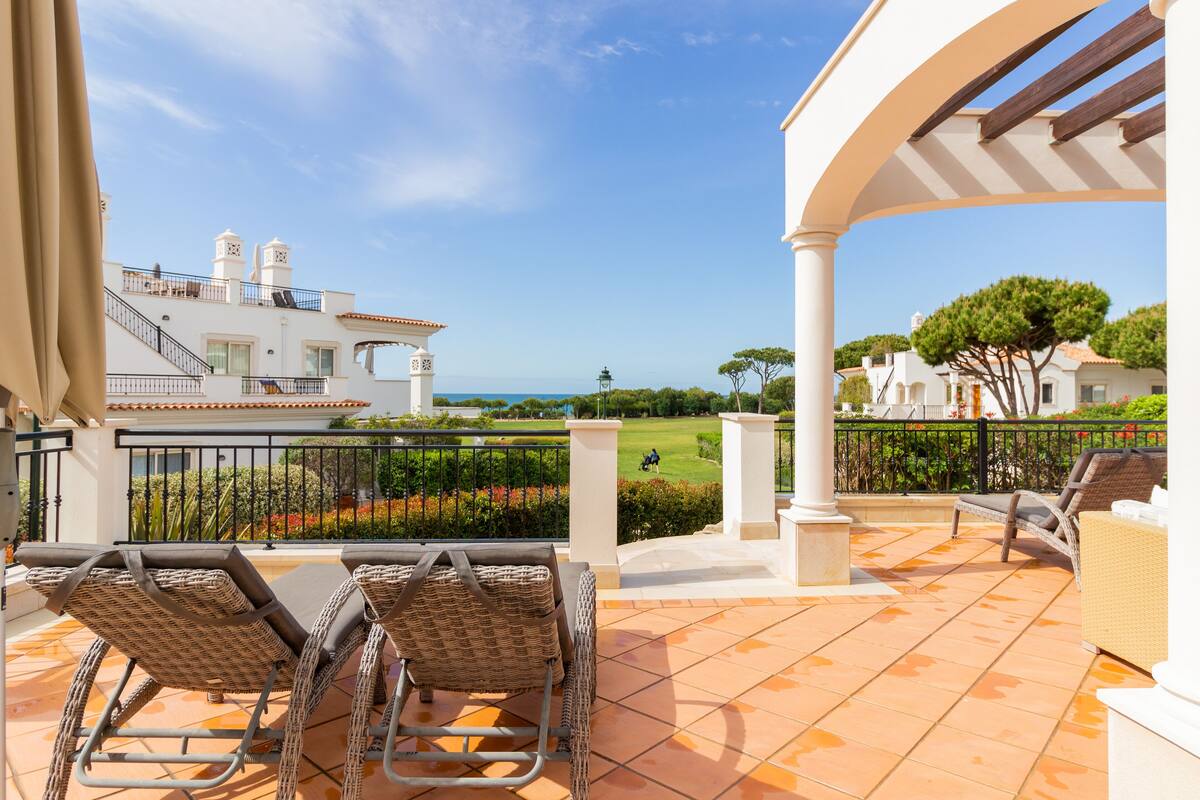 A terrace with wicker chairs, a pergola, and a view of a golf course and ocean.