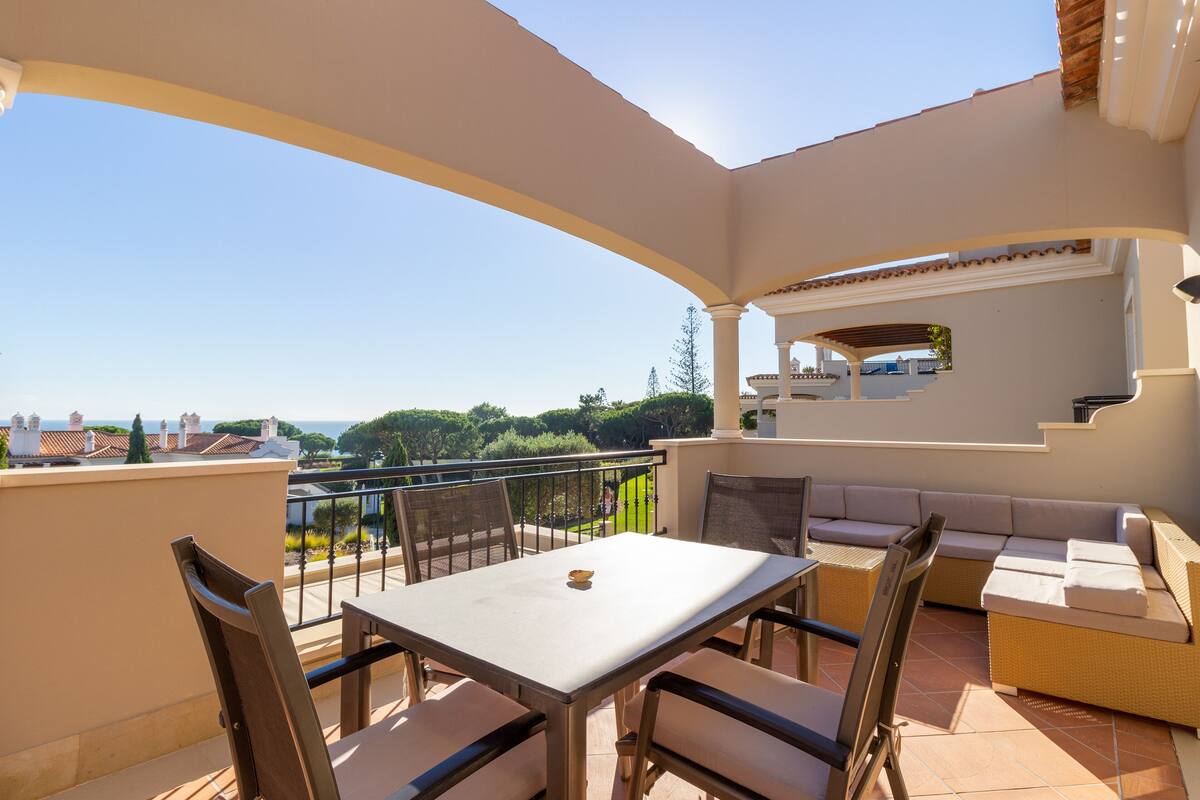 A balcony with a table and chairs, a sofa, and a view of the sea and buildings.