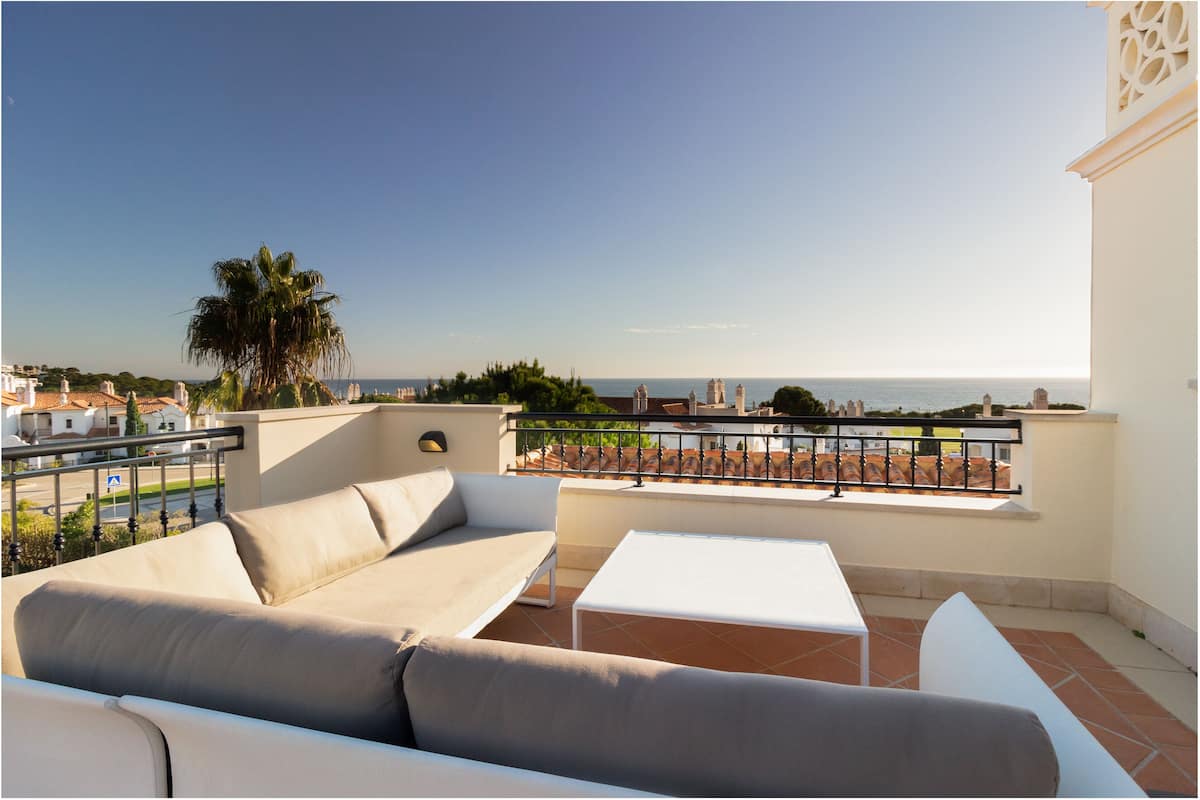 A balcony with a beige sofa, a white table, and a view of the sea and buildings.