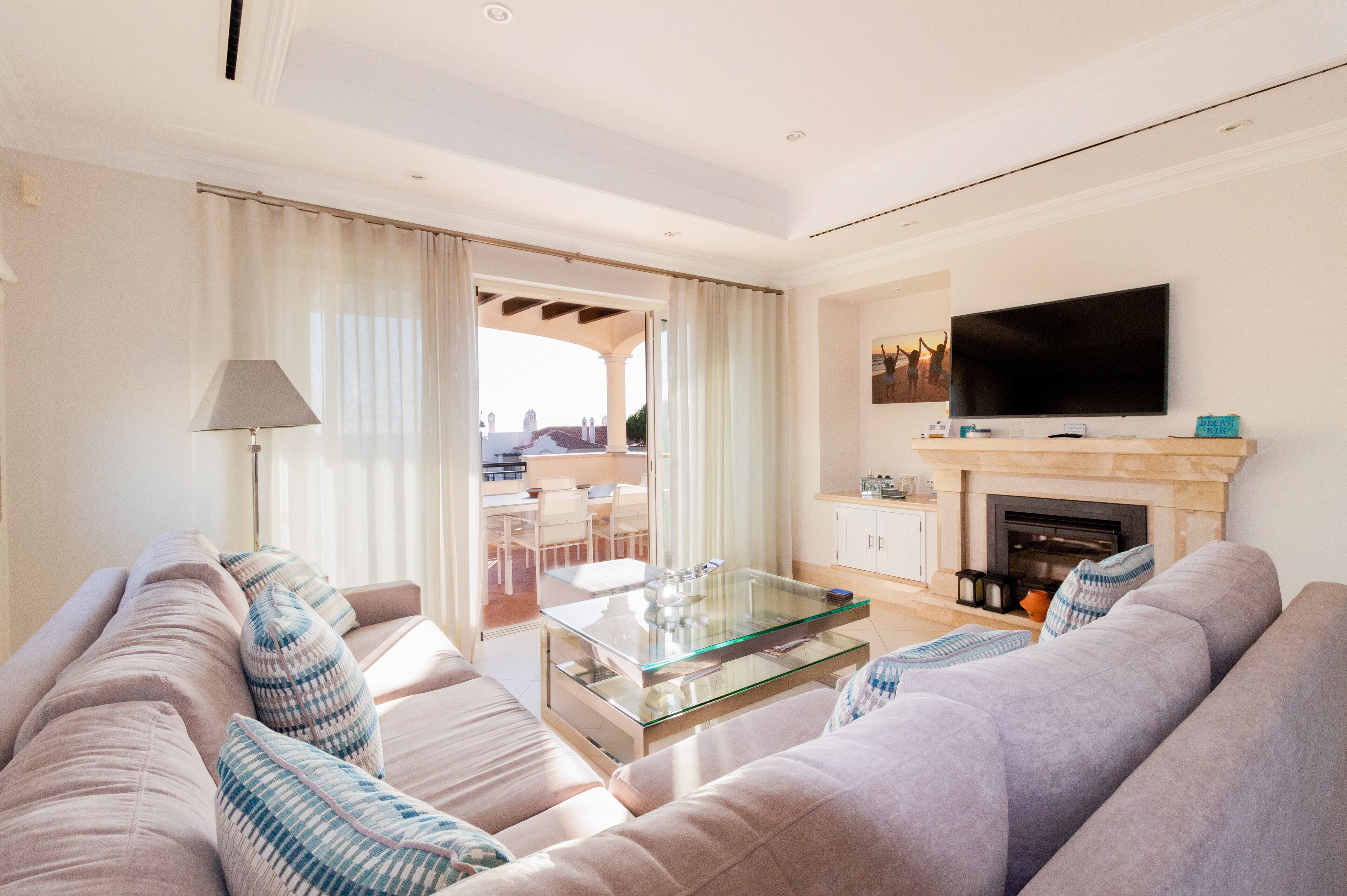 A modern living room with a glass coffee table, a flat-screen TV mounted above a fireplace, and a sofa with patterned cushions.