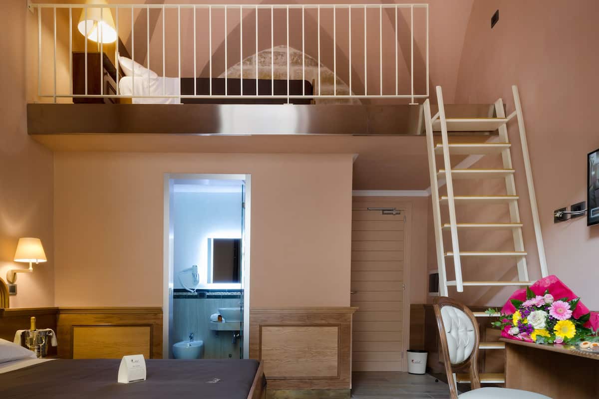 A bedroom with a loft bed, a desk with a chair, a staircase, and a bathroom visible through an open door.