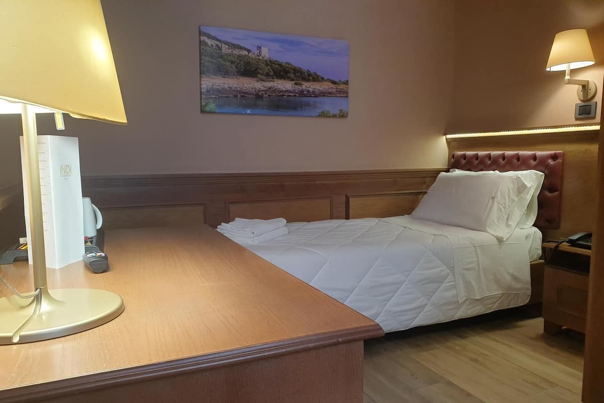 A hotel room with a bed, a desk with a lamp, a painting on the wall, and a nightstand with a lamp.